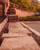 A series of concrete blocks arranged in a row, bordering a pathway or parking area. The setting includes a wooden fence to the left and lush greenery in the background, with a few parked cars visible. The image captures a warm, late afternoon or early evening ambiance.