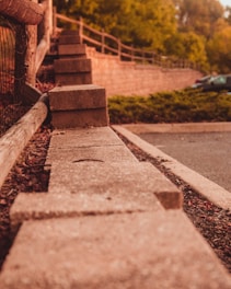 A series of concrete blocks arranged in a row, bordering a pathway or parking area. The setting includes a wooden fence to the left and lush greenery in the background, with a few parked cars visible. The image captures a warm, late afternoon or early evening ambiance.