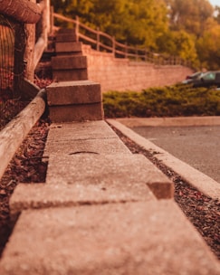 A series of concrete blocks arranged in a row, bordering a pathway or parking area. The setting includes a wooden fence to the left and lush greenery in the background, with a few parked cars visible. The image captures a warm, late afternoon or early evening ambiance.