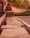 A series of concrete blocks arranged in a row, bordering a pathway or parking area. The setting includes a wooden fence to the left and lush greenery in the background, with a few parked cars visible. The image captures a warm, late afternoon or early evening ambiance.