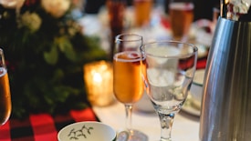 A table is elegantly set with a selection of glassware, including flutes filled with a golden beverage and empty wine glasses. A metal ice bucket is visible to the right, and there is a festive floral arrangement in the background. Warm lighting adds to the cozy and inviting atmosphere.