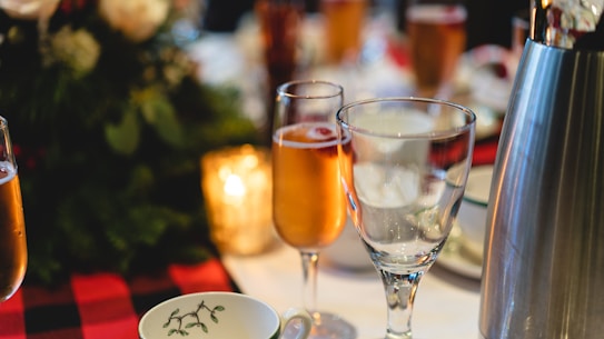 A table is elegantly set with a selection of glassware, including flutes filled with a golden beverage and empty wine glasses. A metal ice bucket is visible to the right, and there is a festive floral arrangement in the background. Warm lighting adds to the cozy and inviting atmosphere.