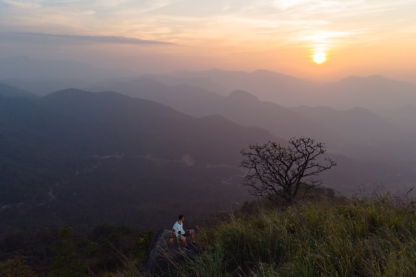 A traveler sitting on a cliff edge, gazing out over rolling hills bathed in soft morning light.