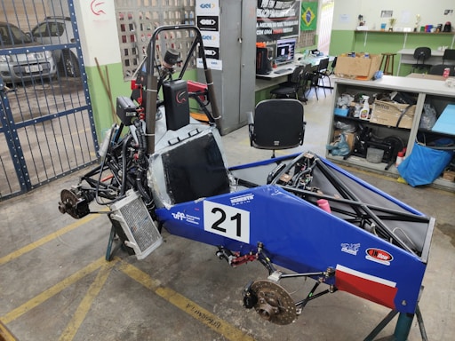 A small, open-frame race car chassis is in a workshop with various tools and equipment scattered around. The car frame is blue with a number '21' displayed on the side. There are exposed components such as wheels, suspension, and a steering mechanism. The workshop has a concrete floor with yellow markings, a metal gate, and shelves holding equipment and supplies.