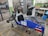 A small, open-frame race car chassis is in a workshop with various tools and equipment scattered around. The car frame is blue with a number '21' displayed on the side. There are exposed components such as wheels, suspension, and a steering mechanism. The workshop has a concrete floor with yellow markings, a metal gate, and shelves holding equipment and supplies.