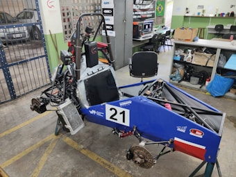 A small, open-frame race car chassis is in a workshop with various tools and equipment scattered around. The car frame is blue with a number '21' displayed on the side. There are exposed components such as wheels, suspension, and a steering mechanism. The workshop has a concrete floor with yellow markings, a metal gate, and shelves holding equipment and supplies.