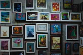 A wall decorated with a variety of framed paintings featuring nature, animals, and floral themes. The artworks include images of rabbits, birds, flowers, a swan on a lake, a forest path in winter, and a girl in a green dress. The arrangement is eclectic, with some paintings hung horizontally and others vertically. The center showcases a framed writing in a foreign script.