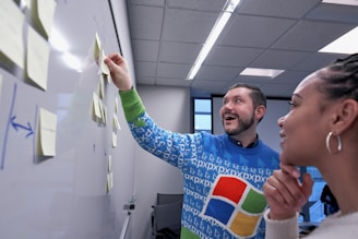 A man and a woman are collaborating in a modern office setting. The man, wearing a colorful sweater with a recognizable tech logo, is enthusiastically placing sticky notes on a whiteboard wall. The woman looks on thoughtfully. The room has a ceiling with fluorescent lighting, and there's a window in the background.