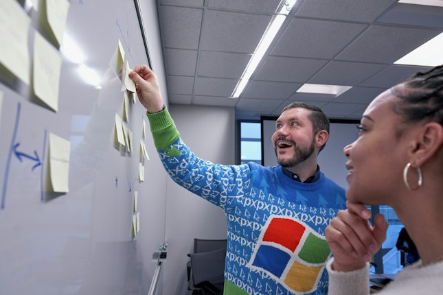 A man and a woman are collaborating in a modern office setting. The man, wearing a colorful sweater with a recognizable tech logo, is enthusiastically placing sticky notes on a whiteboard wall. The woman looks on thoughtfully. The room has a ceiling with fluorescent lighting, and there's a window in the background.