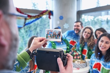 A group of people gathered indoors, wearing festive sweaters and smiling, while one person holds up a Polaroid camera and takes a photo of them. The photo shows the group in the same pose, around a table with holiday decorations, indicating a holiday or celebration scene.