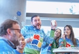 A group of people wearing festive sweaters with colorful designs are enjoying a holiday celebration. One person is holding a cup with a striped straw and appears to be making a playful face, while the others are smiling and eating cookies. Snowflake decorations are visible on a wall in the background.