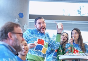A group of people wearing festive sweaters with colorful designs are enjoying a holiday celebration. One person is holding a cup with a striped straw and appears to be making a playful face, while the others are smiling and eating cookies. Snowflake decorations are visible on a wall in the background.