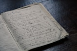 A vintage notebook lies open on a dark wooden surface, filled with cursive handwriting and numerous cross-outs. The paper appears aged, hinting at historical records or personal notes.