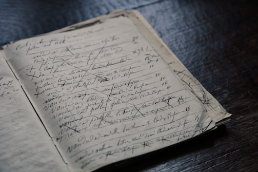 A vintage notebook lies open on a dark wooden surface, filled with cursive handwriting and numerous cross-outs. The paper appears aged, hinting at historical records or personal notes.