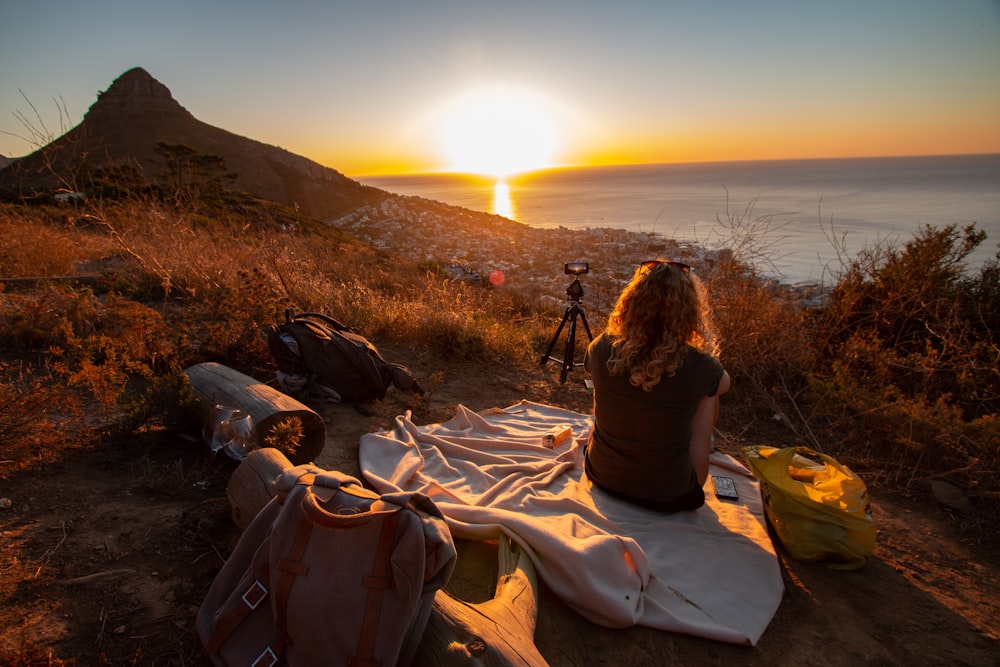 Mujer sentada en la manta cerca del mar y de la montaña durante el amanecer