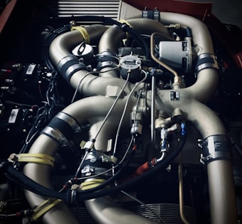 Complex metallic piping system with various hoses and clamps, connected to mechanical components, suggesting an engine or machinery. The arrangement includes multiple curved pipes and wires.