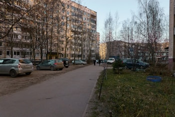 A residential area with several parked cars on a dirt path next to high-rise apartment buildings. Leafless trees are scattered throughout, and a few people can be seen walking along the sidewalk. The grass appears sparse and the buildings are bathed in a warm light.