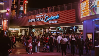 A bustling nighttime scene at the entrance to Universal Orlando Resort. The area is filled with people walking in various directions. Bright lights and signage, such as Starbucks Coffee, Seafood, and a large welcome sign, illuminate the space. The vibrant surroundings, colorful advertisements, and storefronts create a lively atmosphere.