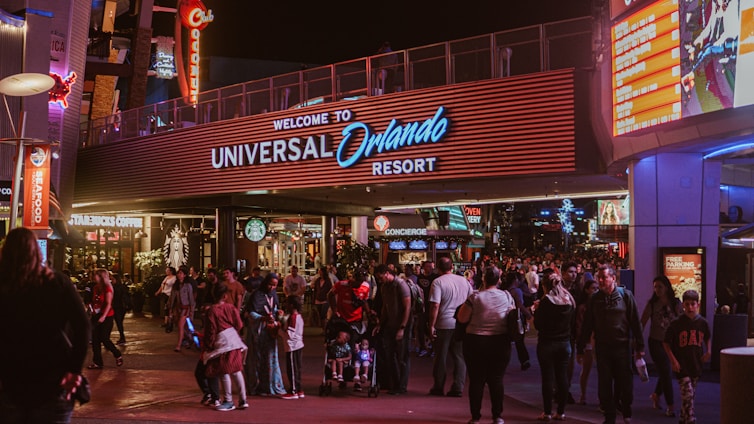 A bustling nighttime scene at the entrance to Universal Orlando Resort. The area is filled with people walking in various directions. Bright lights and signage, such as Starbucks Coffee, Seafood, and a large welcome sign, illuminate the space. The vibrant surroundings, colorful advertisements, and storefronts create a lively atmosphere.