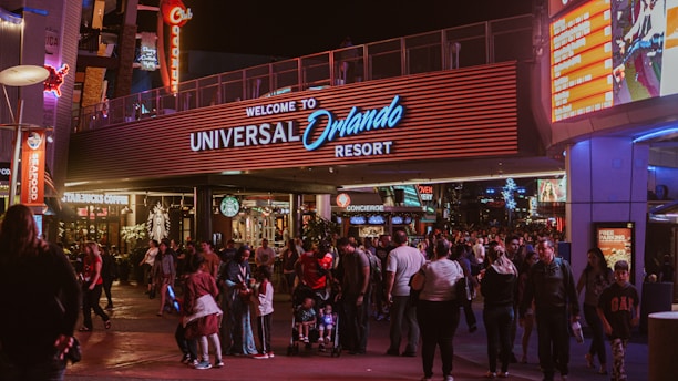 A bustling nighttime scene at the entrance to Universal Orlando Resort. The area is filled with people walking in various directions. Bright lights and signage, such as Starbucks Coffee, Seafood, and a large welcome sign, illuminate the space. The vibrant surroundings, colorful advertisements, and storefronts create a lively atmosphere.