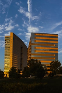 Two modern office buildings with reflective glass windows are set against a backdrop of a bright blue sky with wispy clouds. The buildings have a mix of vertical and horizontal lines, with one of them catching the golden hues of the setting sun. Trees and greenery populate the foreground, adding a natural element to the urban scene.
