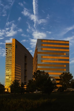 Two modern office buildings with reflective glass windows are set against a backdrop of a bright blue sky with wispy clouds. The buildings have a mix of vertical and horizontal lines, with one of them catching the golden hues of the setting sun. Trees and greenery populate the foreground, adding a natural element to the urban scene.