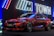 A sleek red BMW M2 car is displayed under vibrant neon signage, featuring the letters 'M Town' in a futuristic setting. The car's polished surface reflects the neon lights, highlighting its aerodynamic design. It is positioned in a dimly lit area, likely at an auto show or promotional event.