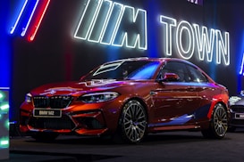 A sleek red BMW M2 car is displayed under vibrant neon signage, featuring the letters 'M Town' in a futuristic setting. The car's polished surface reflects the neon lights, highlighting its aerodynamic design. It is positioned in a dimly lit area, likely at an auto show or promotional event.