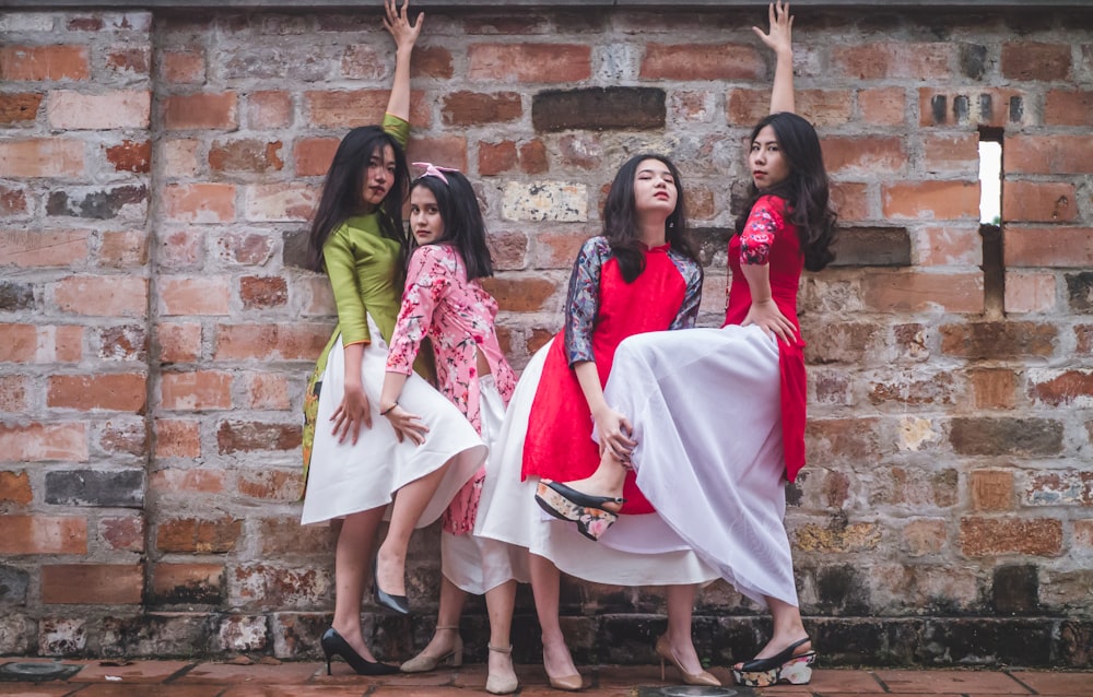 four women in assorted-color dresses standing beside red brick wall