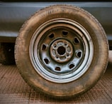 Close-up view of a vehicle wheel with a Bridgestone tire mounted on it, featuring a steel rim. The surface underneath appears textured, possibly suggesting an industrial or outdoor environment.