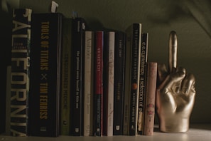 A collection of books neatly arranged on a shelf. The titles visible include 'Tools of Titans' and 'The Consciousness Medicine'. Alongside the books is a decorative golden hand sculpture with the middle finger raised.