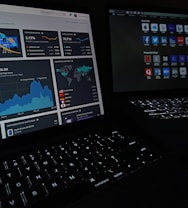 Two laptops display dark-themed screens; one shows analytical graphs and statistics, including conversion rates, store sessions, and geographic distribution. The other screen displays a grid of web application icons. The scene is dimly lit, emphasizing the glow from the screens and the softly illuminated keyboards.
