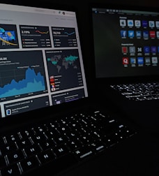 Two laptops display dark-themed screens; one shows analytical graphs and statistics, including conversion rates, store sessions, and geographic distribution. The other screen displays a grid of web application icons. The scene is dimly lit, emphasizing the glow from the screens and the softly illuminated keyboards.