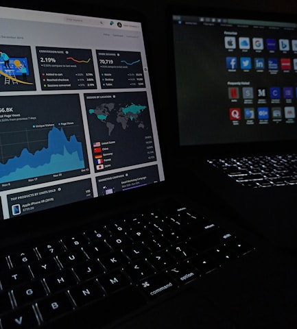 Two laptops display dark-themed screens; one shows analytical graphs and statistics, including conversion rates, store sessions, and geographic distribution. The other screen displays a grid of web application icons. The scene is dimly lit, emphasizing the glow from the screens and the softly illuminated keyboards.