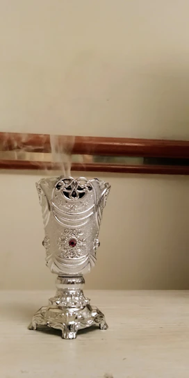 Close-up of an elegant bakhoor burner releasing fragrant smoke against a cream background with gold accents.