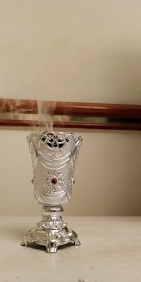 An ornate silver incense holder releases swirling smoke against a plain beige background. The elegant design features intricate patterns and a small red gem embedded at the center. The object is positioned on a flat, light-colored surface, with a wooden railing in the background.