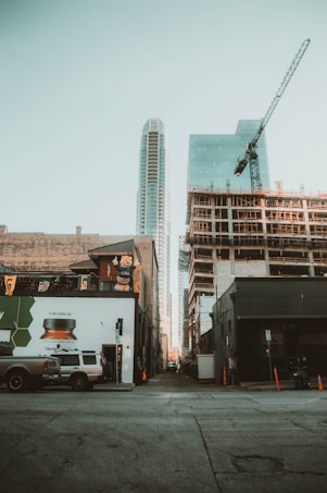 Urban scene with a perspective view down an alley. Tall modern skyscrapers and a construction site with a visible crane rise above older urban buildings. A few parked vehicles and street signs are visible in the foreground.