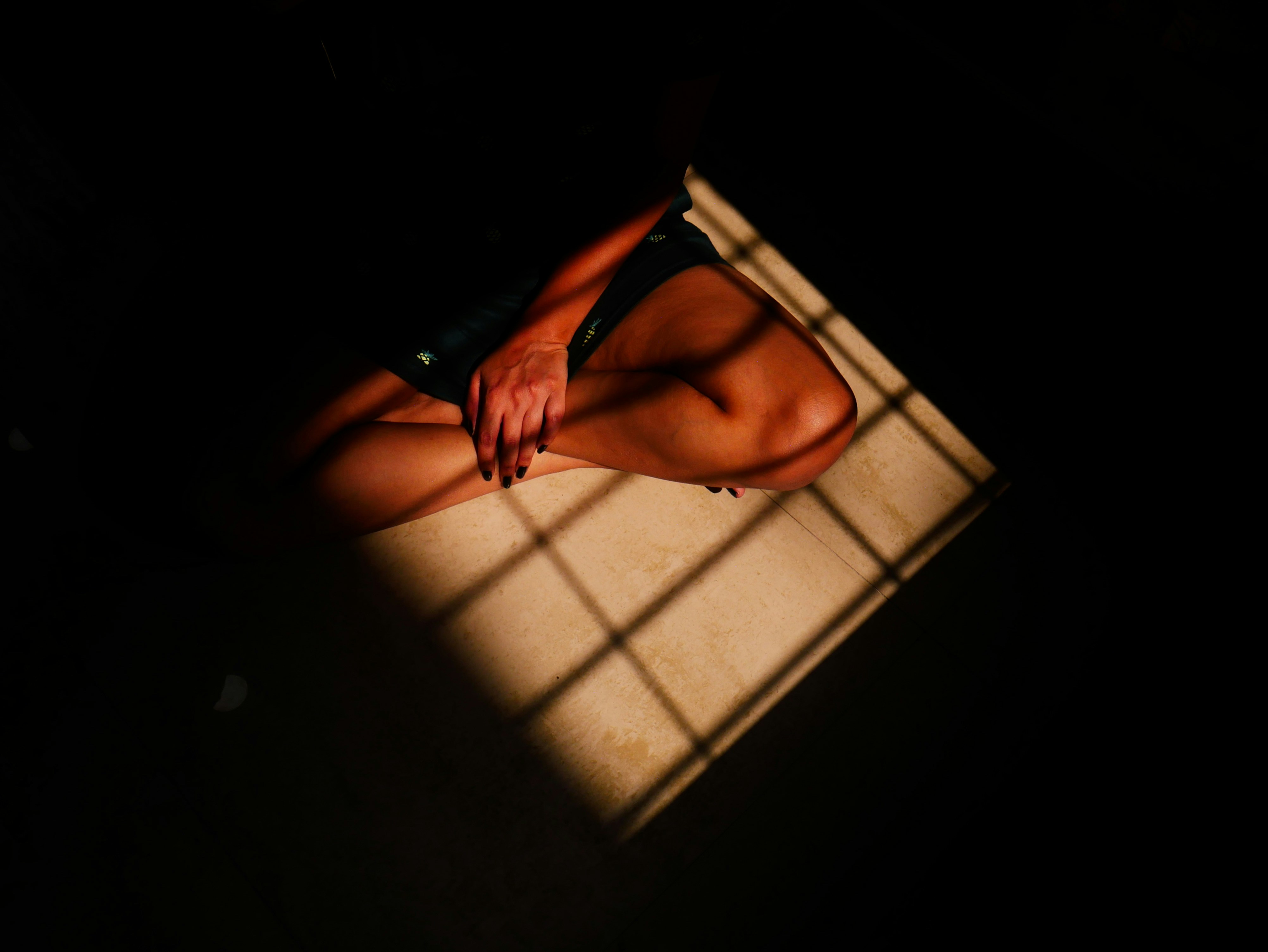 A person with a brown skin tone sitting in shadow on a floor in a square of light crisscrossed with bars.
