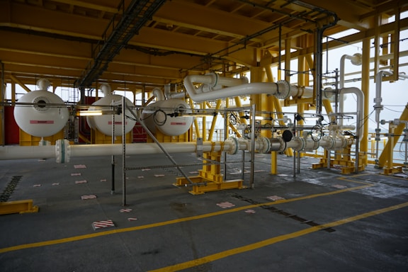 A large industrial setting with intricate piping and machinery connected by large white pipes supported by a yellow metal framework. The environment appears to be part of an offshore platform or processing facility.