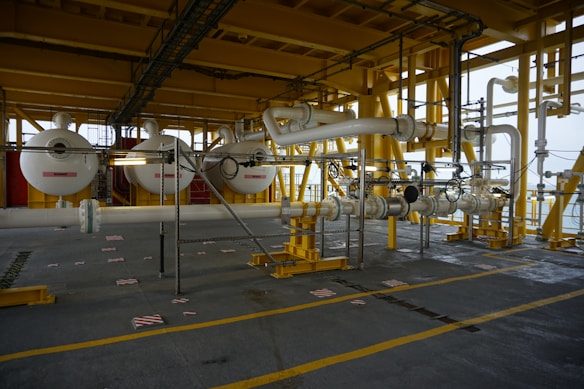 A large industrial setting with intricate piping and machinery connected by large white pipes supported by a yellow metal framework. The environment appears to be part of an offshore platform or processing facility.