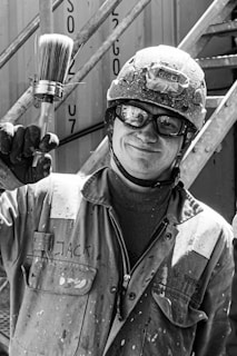 A person wearing a helmet and safety glasses is holding a large paintbrush. The individual is dressed in a work jacket with stains, standing in front of a metal staircase structure. The setting appears industrial with an emphasis on work and safety.