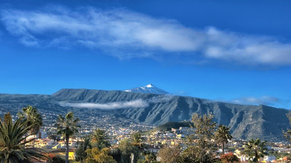 A picturesque landscape featuring a snow-capped mountain in the distance beneath a blue sky with scattered clouds. A vibrant town with colorful buildings is nestled in the valley below, surrounded by lush greenery and palm trees.