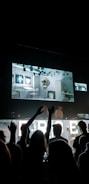 A crowd is gathered in a dark concert or club setting with a large LED screen displaying graphics, including a skull in a brick-walled environment. People in the audience are raising their hands toward the stage.
