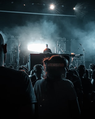 A DJ stands on a stage with bright lights behind them, creating a striking silhouette. The crowd in the foreground watches, partially obscured in shadow, while the stage is filled with smoke and atmospheric lighting. Musical equipment and microphones are visible on the stage.