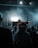 A DJ stands on a stage with bright lights behind them, creating a striking silhouette. The crowd in the foreground watches, partially obscured in shadow, while the stage is filled with smoke and atmospheric lighting. Musical equipment and microphones are visible on the stage.