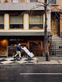 A person wearing a beanie and a dark jacket pushes a hand truck loaded with boxes along a city street. The scene includes a sidewalk and a building facade with large windows and a wooden door. A leafless tree stands in front of the building, and the ground is wet as if it has recently rained.