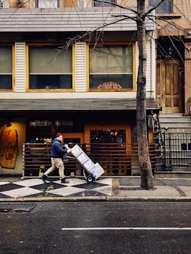 A person wearing a beanie and a dark jacket pushes a hand truck loaded with boxes along a city street. The scene includes a sidewalk and a building facade with large windows and a wooden door. A leafless tree stands in front of the building, and the ground is wet as if it has recently rained.