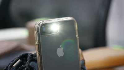 A smartphone is positioned with its rear camera facing upwards, emitting a bright flash of light. The phone is encased in a transparent protective cover. The background is blurred, highlighting the phone as the central focus of the image.