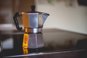 A metal moka pot sits on a smooth, reflective stovetop surface, showcasing its octagonal shape and black handle. The surrounding area is softly blurred, emphasizing the moka pot as the main subject. Its metallic surface reflects light and displays a subtle logo imprinted on the base.