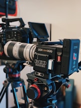 A professional video camera setup is mounted on a tripod. The camera features a large lens and a monitor screen, with various dials and buttons visible on the equipment. Another camera and tripod can be seen in the background.